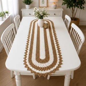 Puede incluir: Un camino de mesa de ganchillo ovalado con bandas concéntricas alternas de hilo blanco y marrón. El camino tiene un borde festoneado y está centrado en una mesa de comedor rectangular blanca. Un jarrón blanco con flores blancas sobre él.