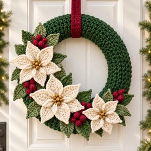 May include: A circular crochet wreath featuring a dark green base, a deep red hanging strap, and three cream-colored poinsettia flowers with gold centers. The arrangement is accented with crocheted green leaves and clusters of small red berries.