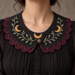 May include: A dark charcoal crochet collar featuring three embroidered golden crescent moons and green leafy vines. The collar has a scalloped deep burgundy border and is fastened at the front with a single round metallic button with a star pattern.