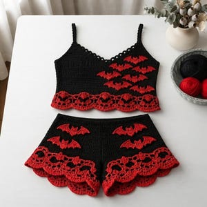 May include: A black crochet two-piece set consisting of a camisole and shorts. The camisole features thin straps, a scalloped neckline, and red bat appliqués. Both pieces have a wide, intricate red crochet lace border with a repeating skull pattern.