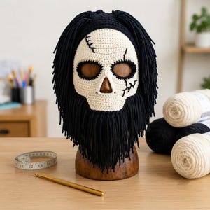 Puede incluir: Una máscara de calavera tejida a ganchillo con rostro color hueso, detalles cosidos en negro y cabello largo de flecos negros. La máscara se exhibe en un soporte de madera, junto a un ganchillo dorado y una cinta métrica de 35 cm.