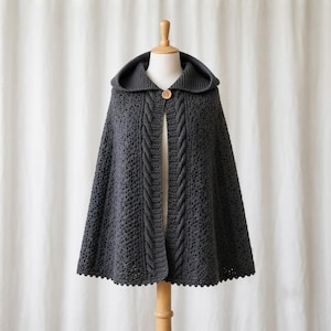 Op de afbeelding: Een donkergrijze cape met capuchon en een ingewikkeld opengewerkt haakpatroon over het hele lichaam. Het kledingstuk is voorzien van een enkele houten knoopsluiting bij de halslijn en verticale kabelbreidetails langs de voorste randen.