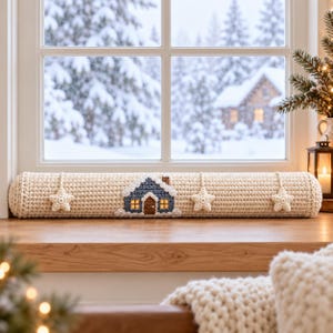 Pode incluir: Um chouriço de porta longo e cilíndrico em croché de cor creme, com uma aplicação central de uma casa azul-escura com telhado coberto de neve e três estrelas de croché cor de creme penduradas na parte frontal do tubo.