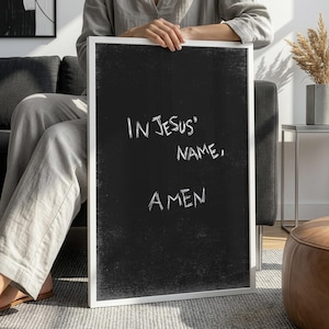 Puede incluir: Impresión artística enmarcada en negro con las palabras blancas escritas a mano "IN JESUS' NAME, AMEN". La obra de arte está sostenida por una persona que viste ropa gris. El fondo incluye un sofá gris, una mesa auxiliar y un puf de cuero marrón.