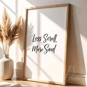 May include: A framed art print with the text "Less Scroll, More Soul" in a stylish script. The artwork is in a light wooden frame and is leaning against a white wall. A vase with dried pampas grass is to the left.