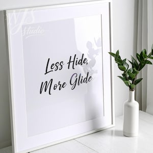 May include: A framed white print with the text "Less Hide, More Glide" in black script. A white vase with green leaves sits to the right of the frame. The image is set against a white background.