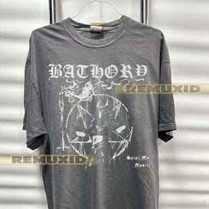 Op de afbeelding: Een grijs t-shirt met de bandnaam "BATHORY" in gotische letters boven een witte afbeelding van een pentagram en een schedel. De tekst "Satan My Master" staat eronder. Het shirt hangt aan een metalen rek.