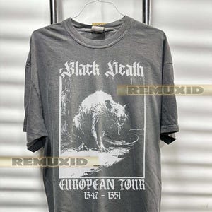 Puede incluir: Camiseta gris con un gráfico blanco de una rata y las palabras "Black Death" y "European Tour 1347-1351". La camiseta está colgada en un perchero de metal.