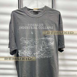 Puede incluir: Camiseta gris oscuro con el texto "VISUALIZE INDUSTRIAL COLLAPSE" sobre un gráfico blanco de un oso en una escena de bosque. La camiseta está colgada en un perchero de metal.