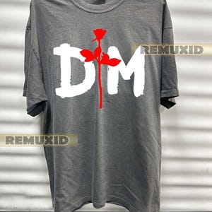 Może przedstawiać: Ciemnoszary t-shirt z krótkim rękawem z dużą białą grafiką liter DM oraz czerwoną łodygą róży umieszczoną pionowo między nimi. Napis REMUXID pojawia się w dwóch miejscach na koszulce, zapisaną małą, prostokątną czcionką.