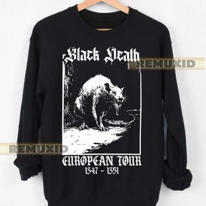 Puede incluir: Sudadera negra con un gráfico blanco de una rata y las palabras "Black Death" y "European Tour 1347-1351". El diseño es una ilustración vintage. La sudadera está colgada en una percha de madera.