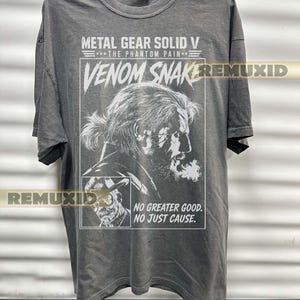 Peut inclure: Un t-shirt gris anthracite à manches courtes avec une impression graphique blanche de Venom Snake de Metal Gear Solid V : The Phantom Pain. Le motif inclut le texte "METAL GEAR SOLID V", "THE PHANTOM PAIN", "VENOM SNAKE" et "NO GREATER GOOD. NO JUST CAUSE."