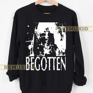 Puede incluir: Sudadera negra con estampado gráfico blanco. El gráfico presenta una imagen distorsionada de una figura y la palabra "BEGOTTEN" en negrita, fuente sans-serif. La sudadera está colgada en una percha de madera.