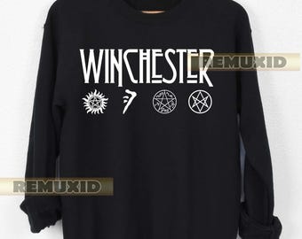 Könnte beinhalten: Schwarzes Sweatshirt mit dem Wort "WINCHESTER" in weißen Großbuchstaben auf der Brust. Darunter befinden sich drei weiße Symbole. Das Sweatshirt hängt an einem Holzkleiderbügel.