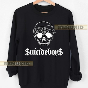 Puede incluir: Sudadera negra con un gráfico de calavera blanca que lleva un pañuelo, con el texto "G59" visible en la frente. Las palabras "$uicideboy$" están impresas en una fuente gótica debajo de la calavera. La sudadera está colgada en una percha de madera.