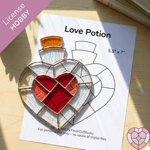 Può includere: Un vetro colorato a forma di cuore con un cuore rosso al centro e vetro bianco e trasparente intorno. Il disegno è su un foglio di carta bianca che dice "Love Potion" e misura 14 cm x 18 cm.