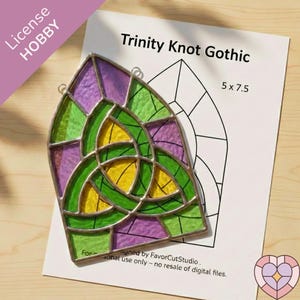May include: A stained glass piece with a Trinity Knot design in green, yellow, and purple, with a silver metal frame. The design is on a white paper that reads "Trinity Knot Gothic" and measures 5 x 7.5 inches.