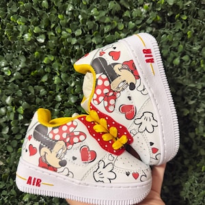 Pu&ograve; includere: Un paio di scarpe da ginnastica bianche con stampa di Minnie Mouse, cuori rossi e pois. Le scarpe hanno lacci gialli, una fodera interna gialla e una suola bianca con la parola AIR stampata in rosso sul lato della zona del tallone.
