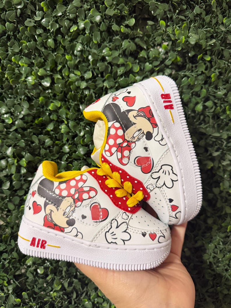 Pu&ograve; includere: Un paio di scarpe da ginnastica bianche con stampa di Minnie Mouse, cuori rossi e pois. Le scarpe hanno lacci gialli, una fodera interna gialla e una suola bianca con la parola AIR stampata in rosso sul lato della zona del tallone.