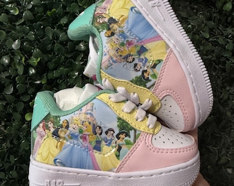 May include: A pair of low-top sneakers featuring a graphic print of various animated princesses and a castle. The shoes have white soles with the word AIR embossed on the side, light pink toe caps, yellow lace panels, and mint green heel tabs.