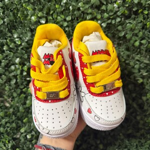 Pu&ograve; includere: Un paio di scarpe da ginnastica bianche con lacci gialli, dettagli rossi a pois bianchi e grafiche di Minnie Mouse. La linguetta riporta la scritta Minnie Mouse e i lacci sono fissati con placchette metalliche dorate con la scritta AFT.