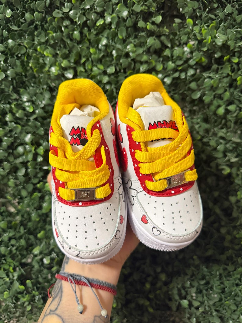 Pu&ograve; includere: Un paio di scarpe da ginnastica bianche con lacci gialli, dettagli rossi a pois bianchi e grafiche di Minnie Mouse. La linguetta riporta la scritta Minnie Mouse e i lacci sono fissati con placchette metalliche dorate con la scritta AFT.