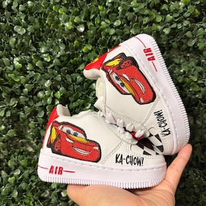 May include: A pair of white sneakers featuring red embroidered race car patches on the sides. The shoes include red heel tabs, white laces, and white soles with the word AIR in red text. The text KA-CHOW! is printed in black on the side panels.