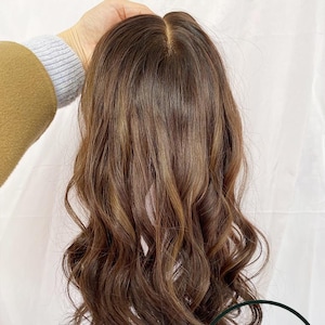May include: A brown wig with long, wavy hair. The wig has a natural-looking parting and is held up by a hand. The hair has highlights and lowlights, giving it dimension. The wig is likely made of synthetic or human hair.