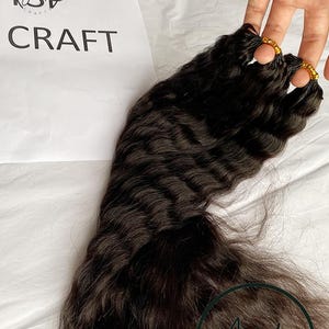 May include: Dark brown wavy hair extensions. The hair is held together with gold rings. The word "CRAFT" is printed on a white paper. The image is taken on a white surface.