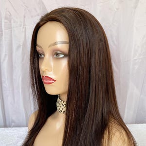 May include: A brown wig with long, straight hair styled on a mannequin head. The mannequin has a fair complexion, red lipstick, and a patterned scarf. The wig is a rich, dark brown colour.