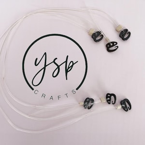 Puede incluir: Cordones de plástico transparente con pequeños ganchos negros y cuentas cuadradas de color claro. Los cordones están dispuestos en forma circular con el texto YSP CRAFTS impreso en un círculo verde oscuro en el centro de la imagen.