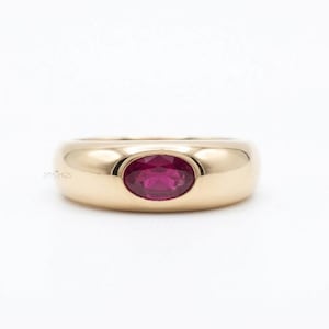 Peut inclure: Une bague anneau en or avec une surface lisse et polie, ornée d'une pierre précieuse rose foncé taille ovale sertie horizontalement au centre. Le métal présente une finition chaude et réfléchissante, et la pierre a une surface facettée.