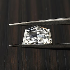 Pode incluir: Uma pedra preciosa transparente e incolor em forma de escudo, segurada por uma pinça de metal. A pedra apresenta um corte geométrico com múltiplas superfícies refletoras e ângulos agudos. O texto ZOSIMOS está impresso em letras pretas.