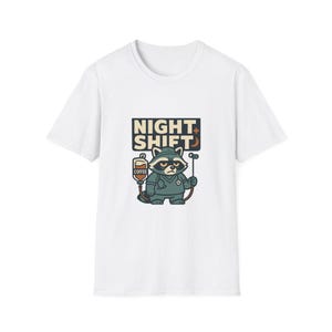 May include: A white short-sleeve t-shirt featuring a graphic of a raccoon dressed in medical scrubs. The raccoon holds an IV bag labeled COFFEE and the text NIGHT SHIFT with a small crescent moon icon above the character in dark blue and tan.