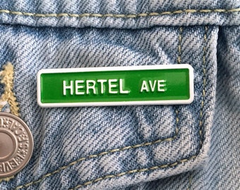 May include: A green and white enamel pin with the text "Hertel Ave" on a denim jacket.