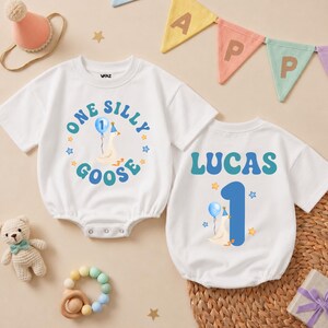 May include: Two white short-sleeved baby bodysuits featuring colorful graphics. One displays a goose with a blue balloon and the text One Silly Goose. The other shows the name Lucas, a large blue number 1, and a goose with a blue balloon.
