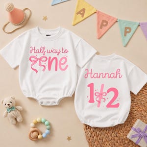May include: Two white short-sleeved baby bodysuits with pink text and graphics. One reads Half way to one with a pink bow, the other reads Hannah 1/2 with a pink bow. Includes a small party hat, a crochet bear, a bead teether, and a banner.