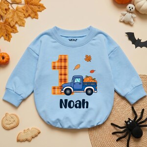 May include: A light blue long-sleeve sweatshirt featuring a large orange plaid number one, a blue pickup truck carrying pumpkins, and the name Noah printed in dark blue. The garment has a crew neckline and ribbed cuffs, with the brand name VFAZ visible.