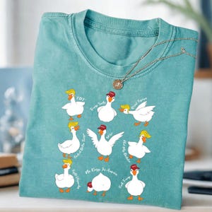 May include: A teal t-shirt with cartoon geese wearing blonde wigs and red hats. The geese have phrases like "Trump Sucks" and "Resist Fascism" written around them. A silver necklace with a pendant is draped over the shirt.