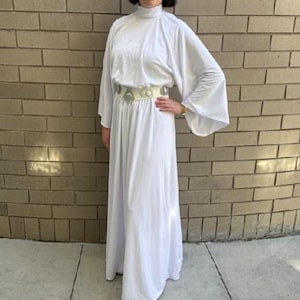 May include: A floor-length white dress with long, wide sleeves and a high neckline. The garment features a metallic silver belt with circular accents at the waist. The fabric is smooth and opaque, draping down to the floor in a simple, straight cut.