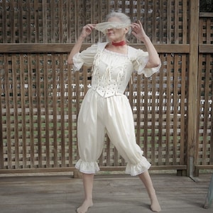 May include: A woman wearing a white lace-trimmed top and matching pants with a red ribbon around her neck. She is holding a white lace veil over her face.