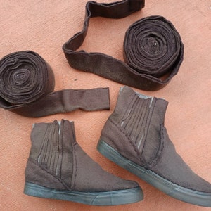 May include: Two brown ankle boots with a brown felt upper and a black rubber sole. The boots are sitting on a brown surface. There are three rolls of brown felt fabric in the background.