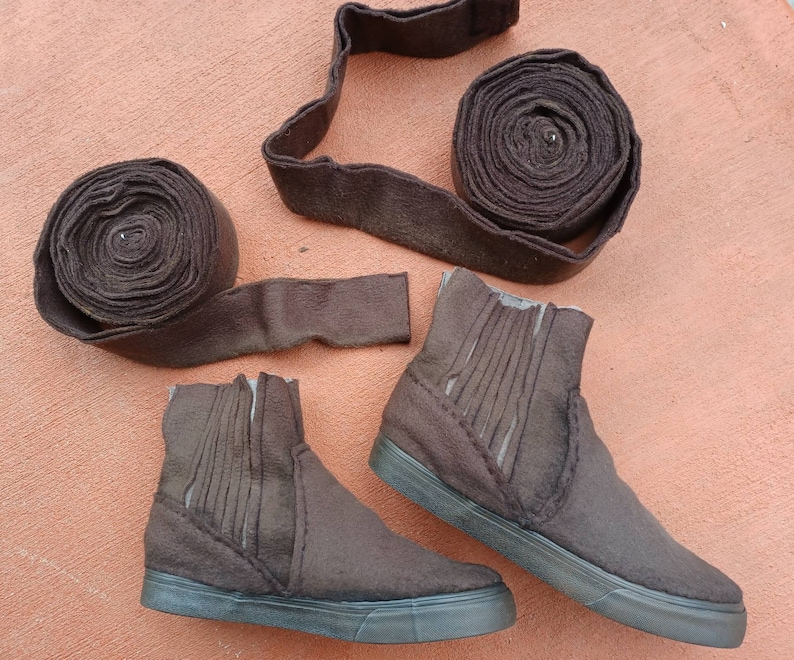 May include: Two brown ankle boots with a brown felt upper and a black rubber sole. The boots are sitting on a brown surface. There are three rolls of brown felt fabric in the background.