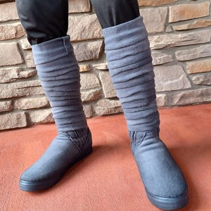 May include: Gray, textured boots with a ribbed design extending up the leg. The boots have a flat sole and are worn with black pants. The boots appear to be made of a soft, felt-like material.