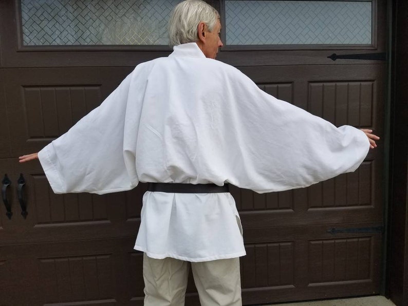 May include: A white tunic with long sleeves and a black belt. The tunic is made of a soft, flowing fabric and has a simple, elegant design.