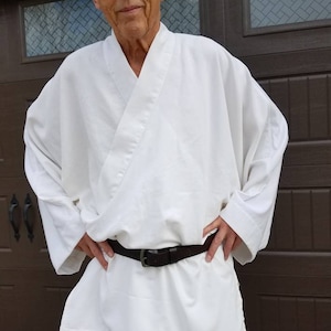May include: A white robe with a brown belt. The robe has long sleeves and a wide collar. The person wearing the robe is standing in front of a brown door.