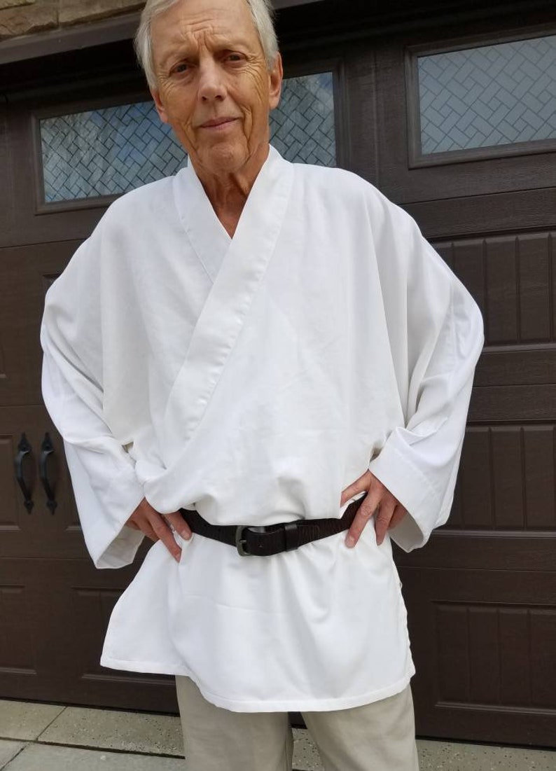 May include: A white robe with a brown belt. The robe has long sleeves and a wide collar. The person wearing the robe is standing in front of a brown door.