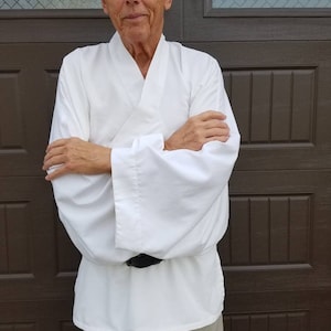 May include: A man wearing a white, long-sleeved shirt with a wide collar and a black belt. He is standing in front of a brown wooden door.