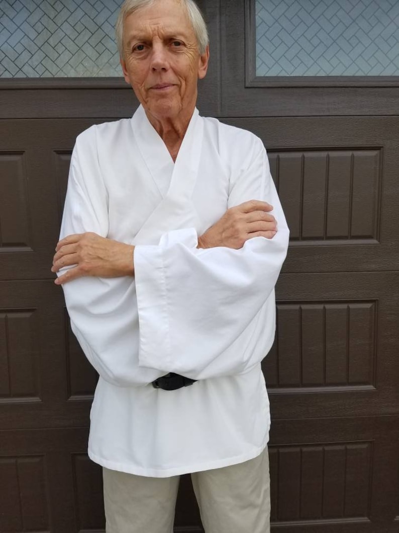 May include: A man wearing a white, long-sleeved shirt with a wide collar and a black belt. He is standing in front of a brown wooden door.