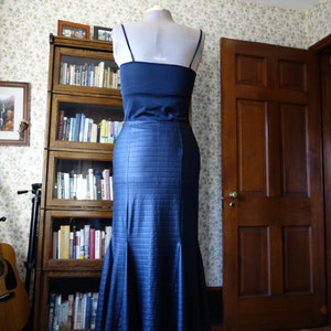 May include: A dark blue mermaid-style skirt with a black lace trim. The skirt is on a mannequin and is displayed in front of a bookcase and a wooden door.